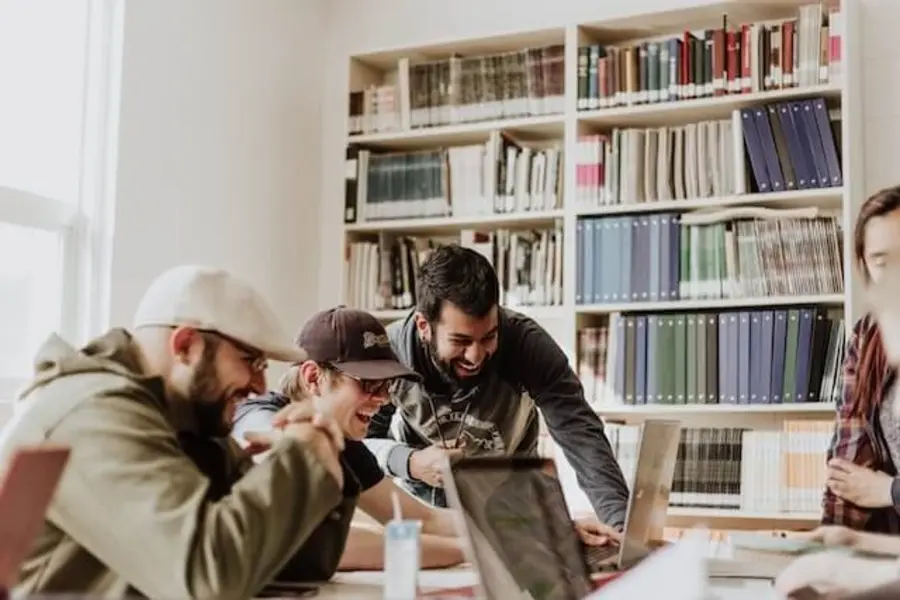Three men are laughing as they look at a laptop inside a room filled with educational books.
